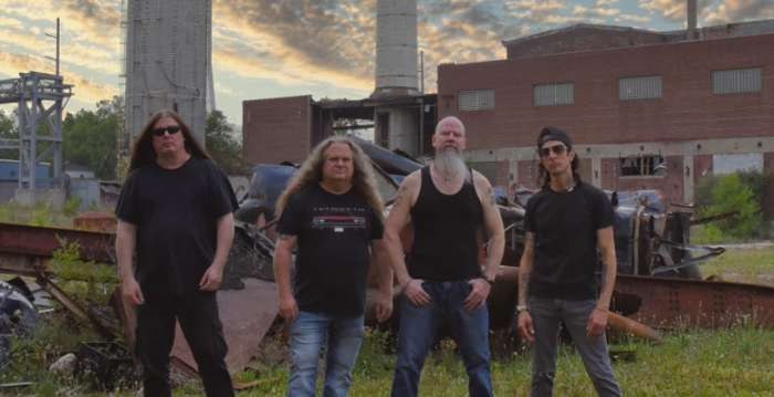 Fotografia de promoció d'Exhorder: els quatre membres del grup drets davant d'una nau industrial abandonada i de ferralla, al capvespre
