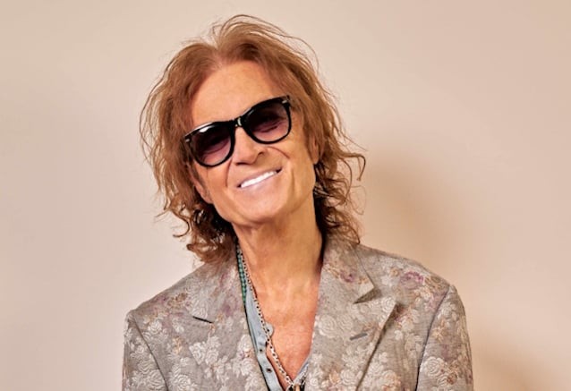 Glenn Hughes somrient, amb ulleres de sol fosques i americana estampada de flors, en una foto de promoció sobre fons clar