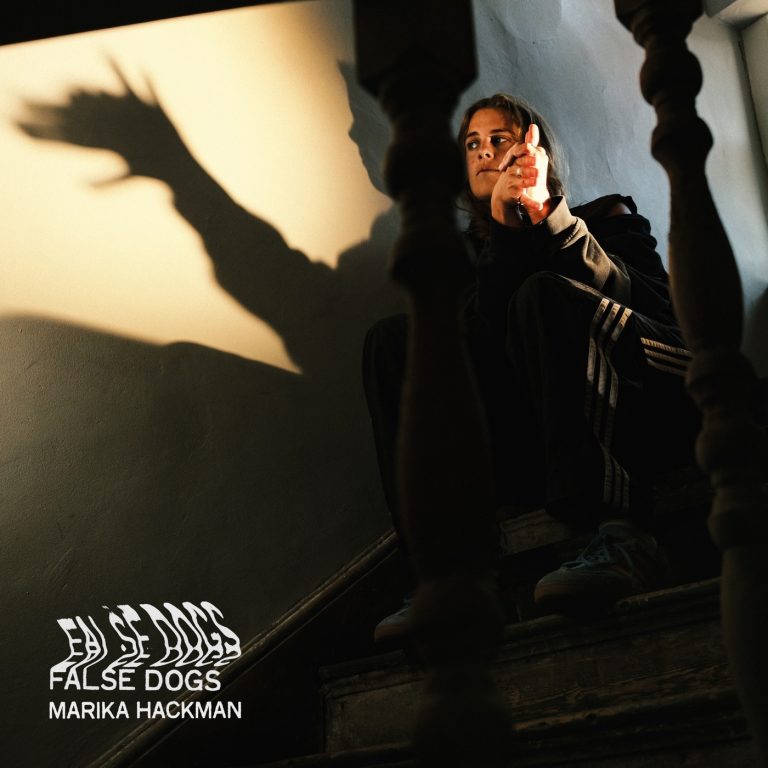 Portada de «False Dogs», de Marika Hackman: la cantant asseguda en una escala fent amb la mà una ombra xinesa de gos damunt la paret