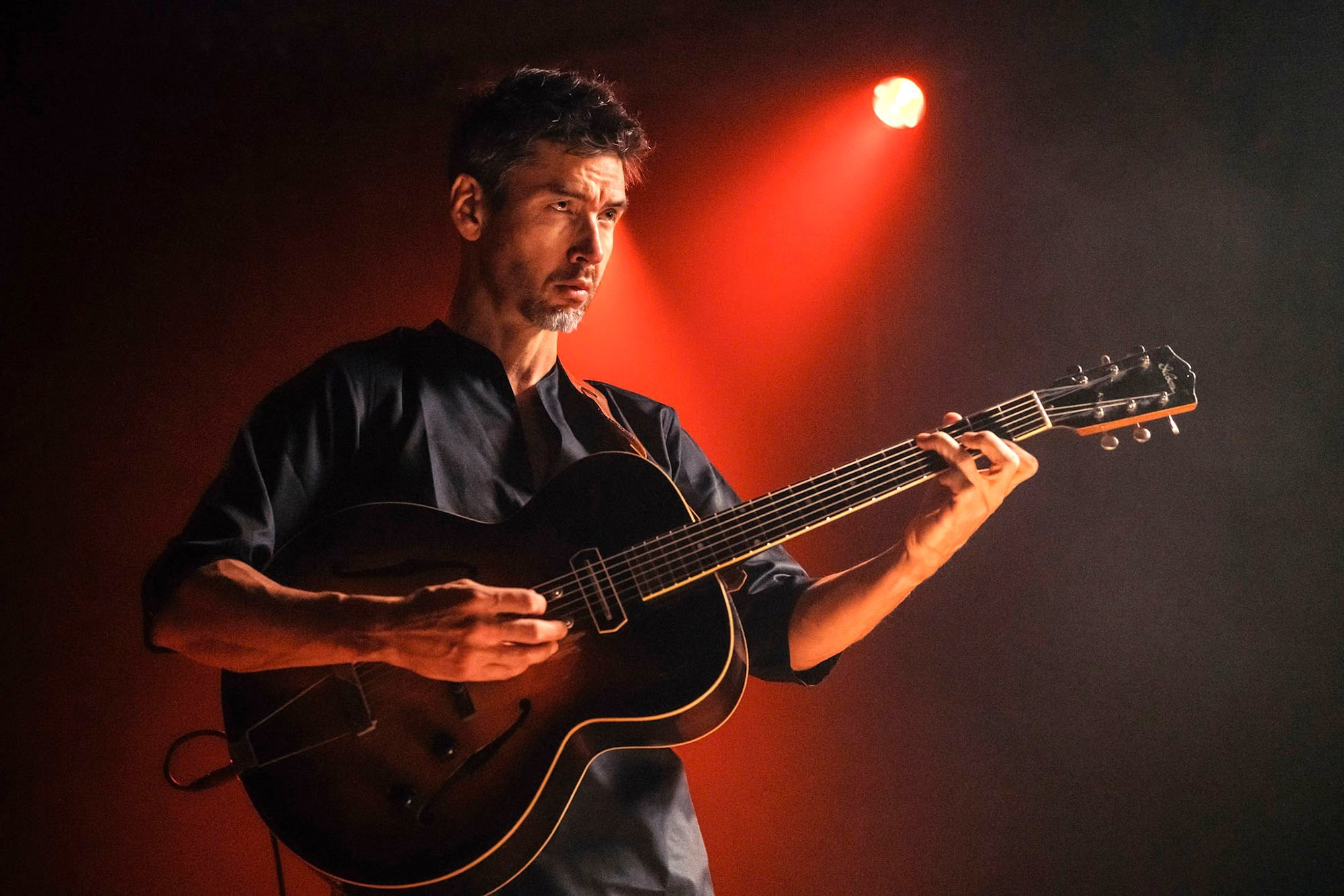 Miles Okazaki assegut sol a l'escenari del Jazz Gallery amb la seva guitarra de tapa arquejada durant el rodatge de «The Monkathon»
