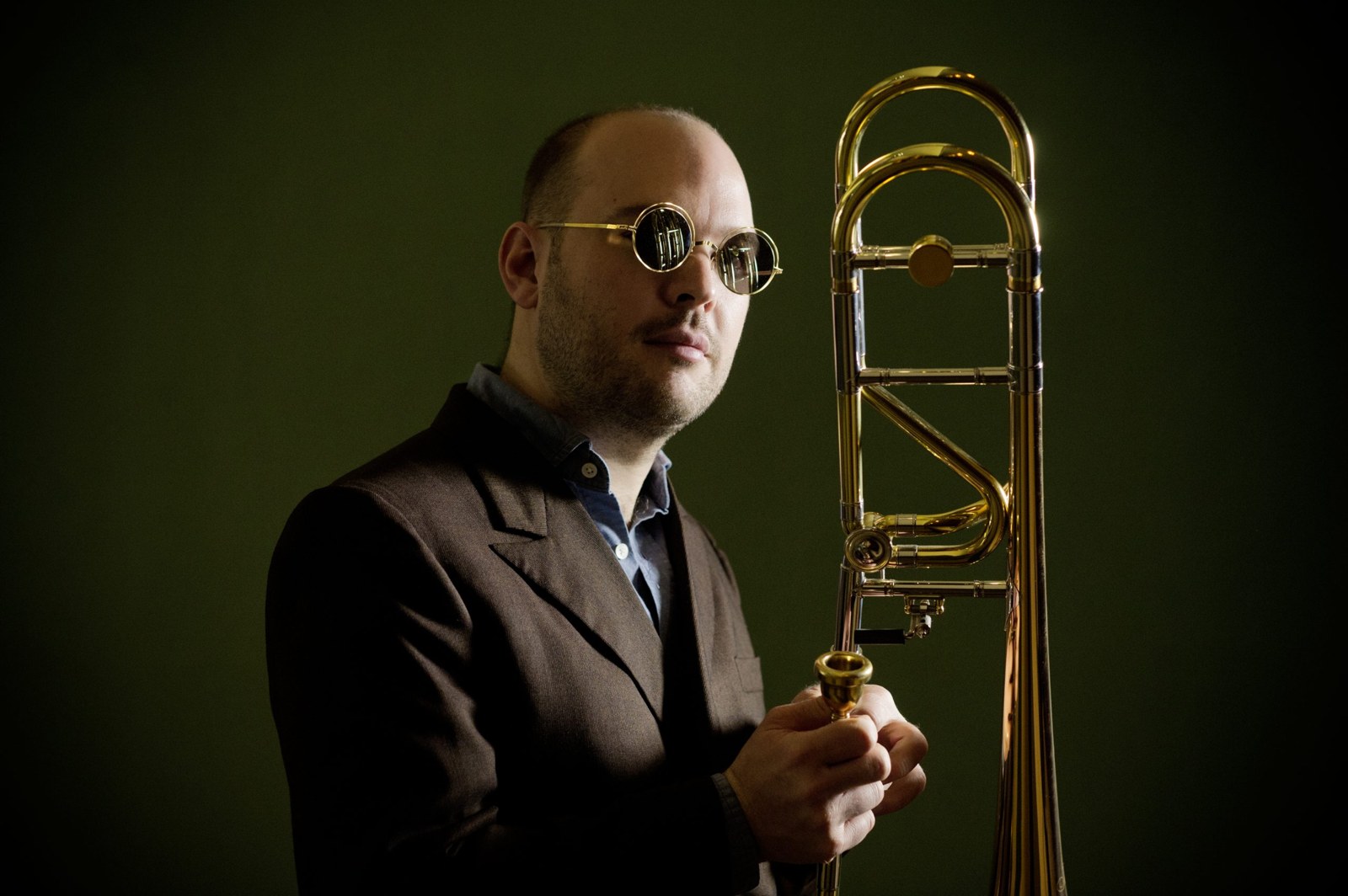 Retrat de Samuel Blaser amb ulleres rodones fosques i americana marró, al costat d'un trombó daurat, sobre fons verd fosc