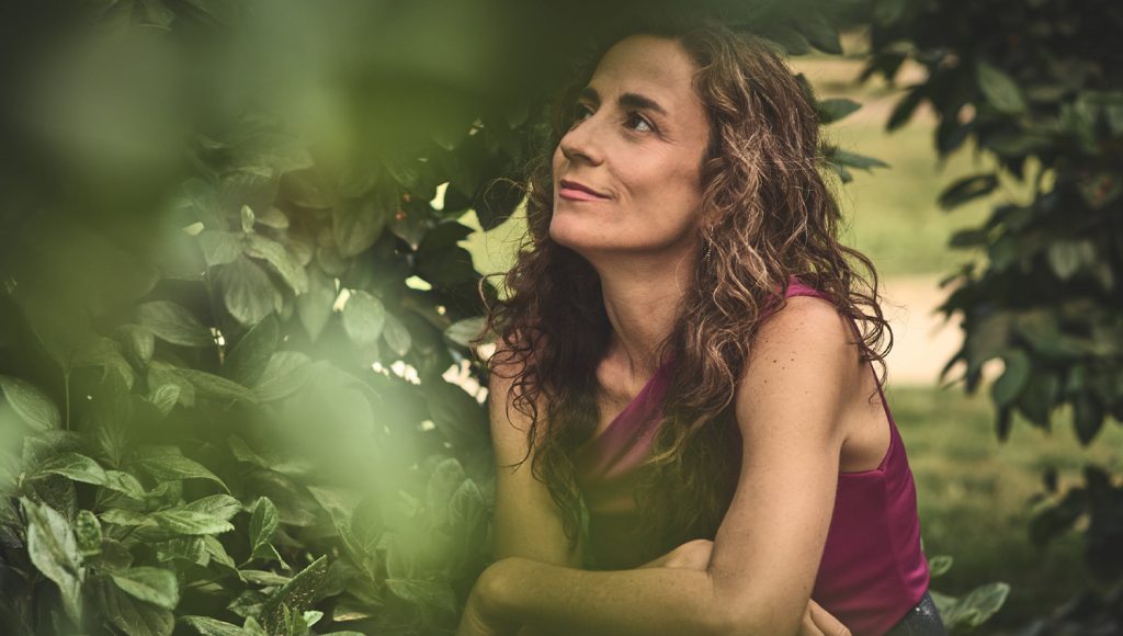 Sofia Ribeiro, amb els cabells llargs i arrissats i una samarreta granat, ajupida entre les fulles d'un arbust i mirant cap amunt
