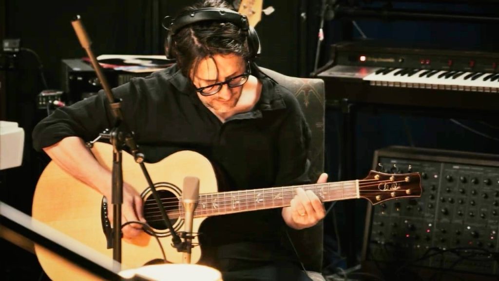 Steven Wilson, amb auriculars i ulleres, toca una guitarra acústica davant d'un micròfon en un estudi amb un teclat i un sintetitzador modular al fons