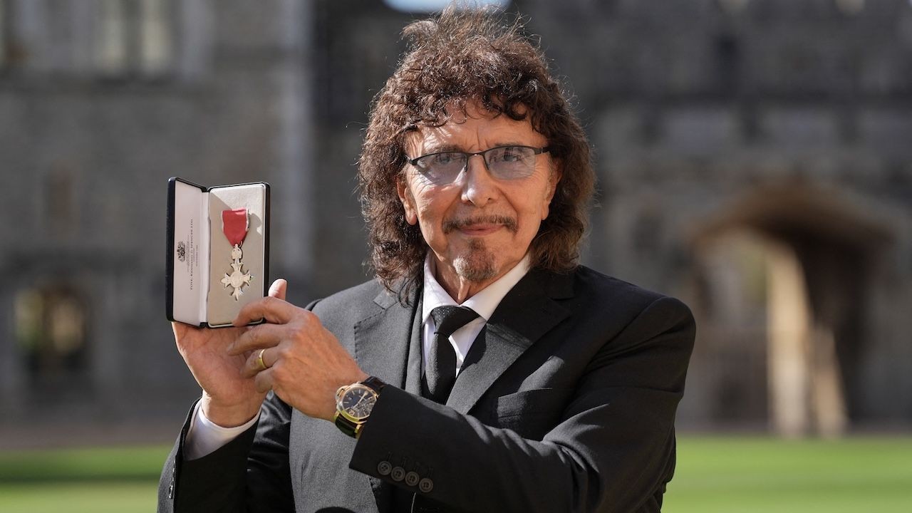 Tony Iommi, amb vestit fosc, aguantant la medalla de la MBE dins de l'estoig obert als jardins del castell de Windsor