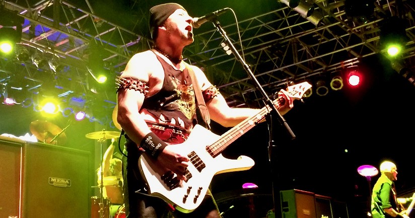 Cronos cantant i tocant el baix blanc damunt d'un escenari il·luminat de verd, amb la bateria de Venom al darrere