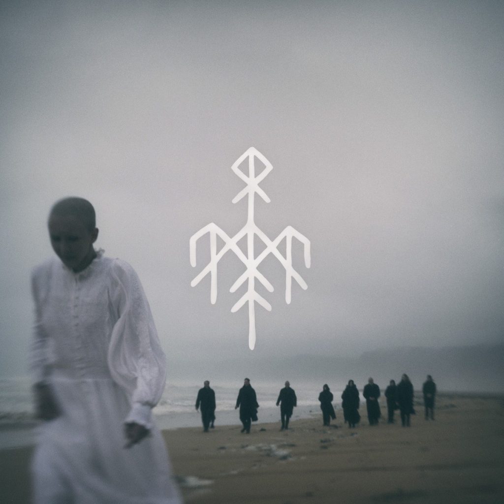 Portada de «Helvegen II», de Wardruna: una dona vestida de blanc camina per una platja boirosa, amb un grup de figures de negre al fons i el símbol rúnic del grup al cel
