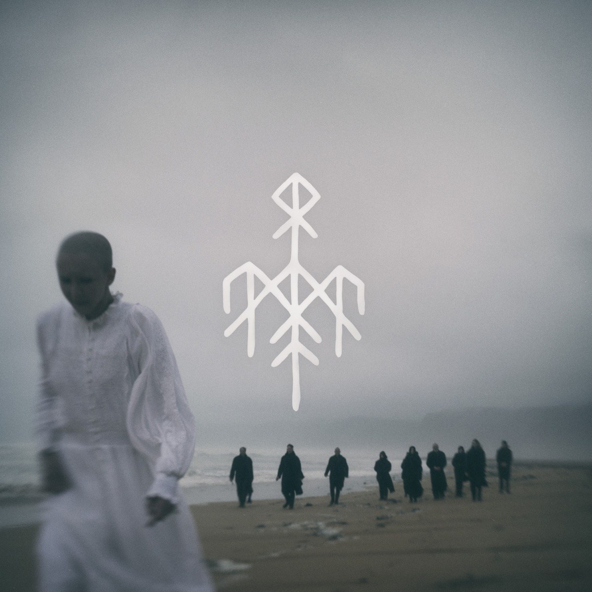 Portada de «Helvegen II», de Wardruna: una dona vestida de blanc camina per una platja boirosa, amb un grup de figures de negre al fons i el símbol rúnic del grup al cel